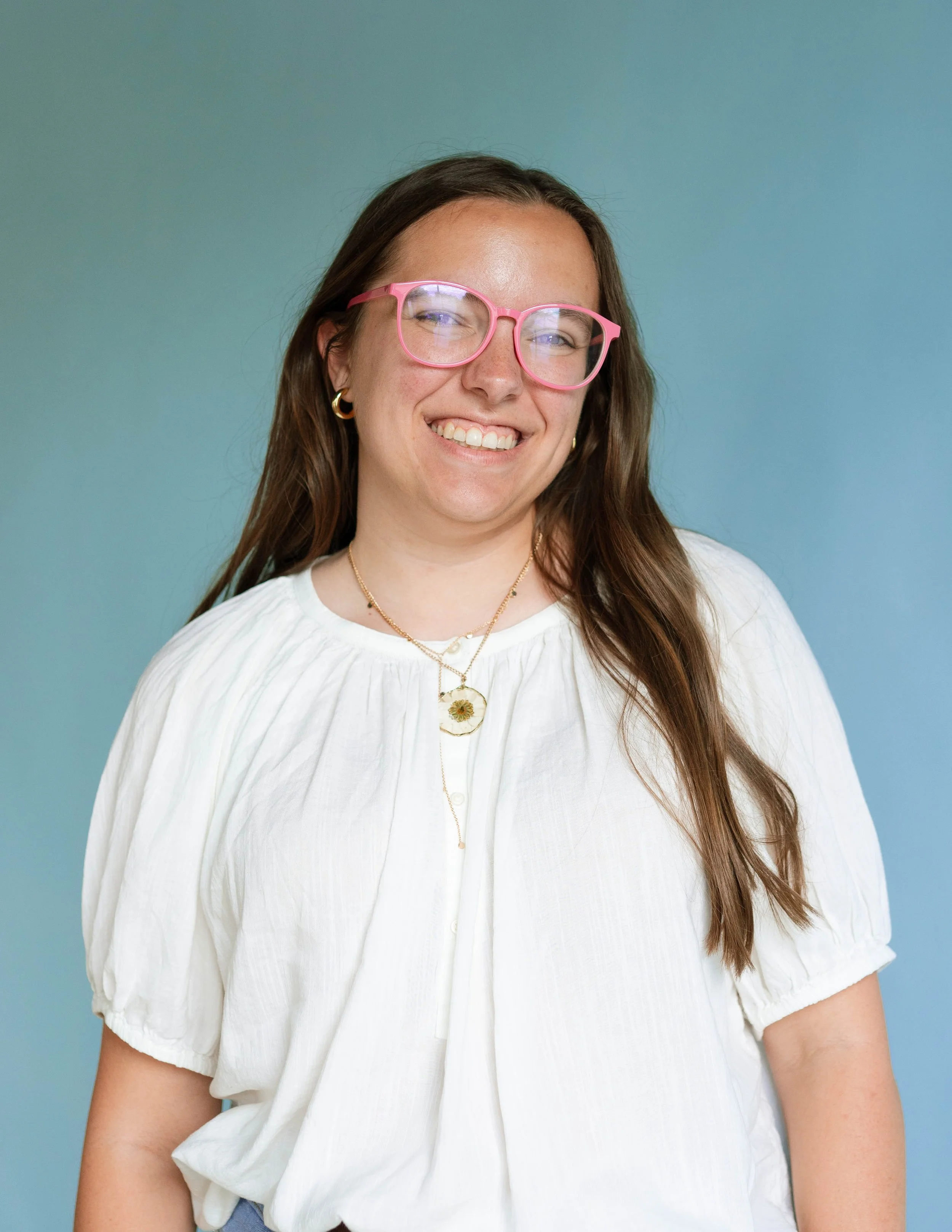 A smiling woman with long brown hair, wearing pink glasses, gold earrings, layered necklaces, and a white blouse, standing against a teal background.