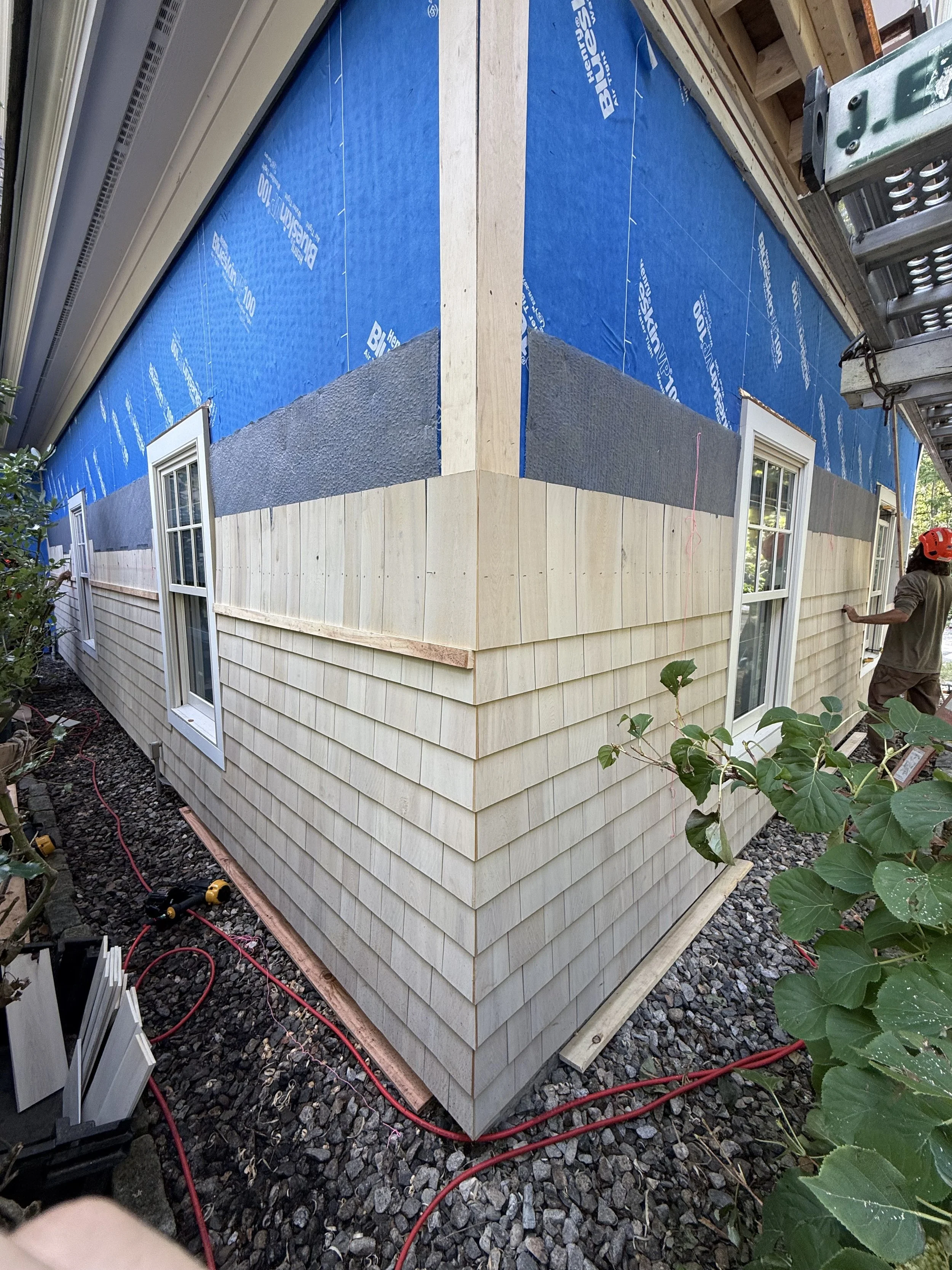 Side view of a house under construction, showing siding and window frames, with tools and red extension cords on the gravel ground.