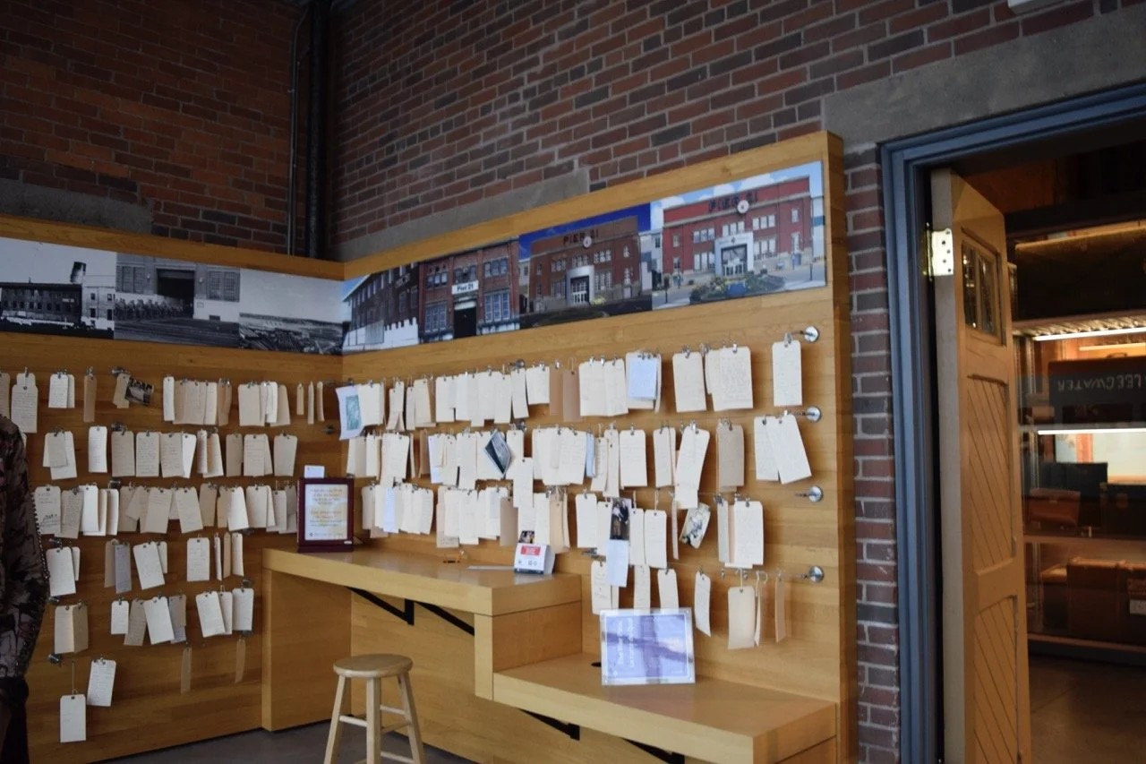 Large paper tags with handwritten notes on them hanging on a wooden wall in an exhibit space.