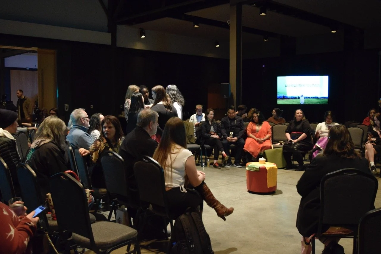 Large group of people sitting in a circle with TV screen in the background.