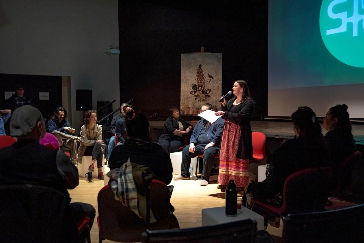 Person with a microphone and piece of paper in hand are standing front of an audience.