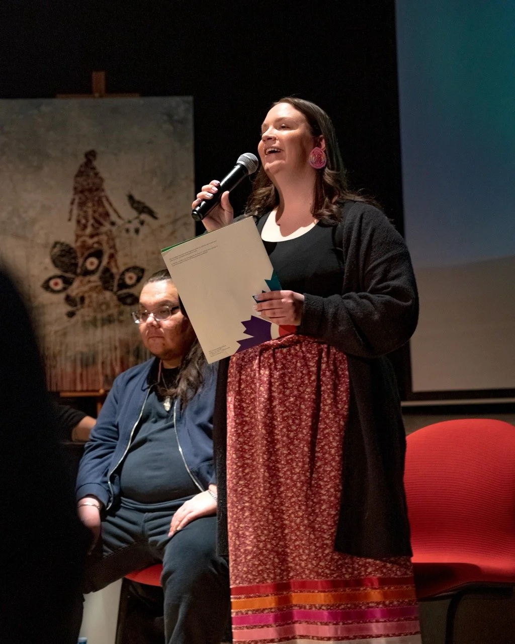 Close up of person with a microphone and piece of paper in hand are standing front of an audience.