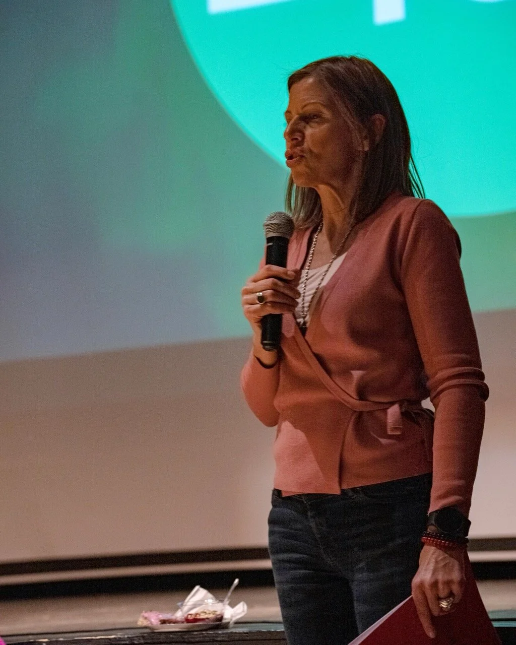 Close up of person with microphone in hand standing front of green background, showing part of the Where We Stand logo.
