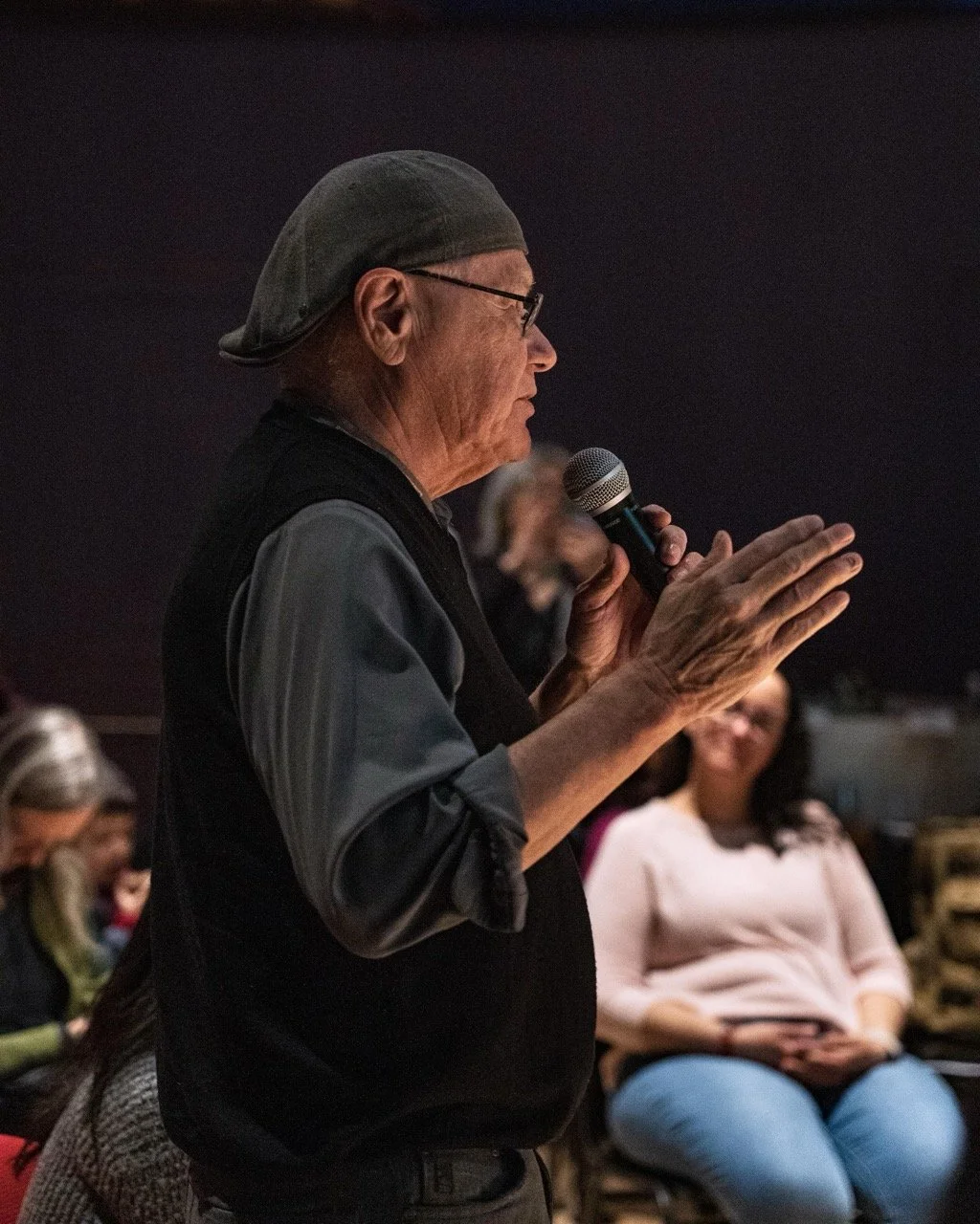 Close up of a person wearing hat and glasses, is standing front of audience with a microphone in hand.