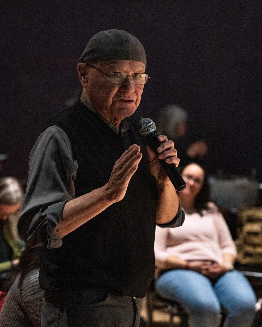 Close up of a person wearing hat and glasses, is standing front of audience with a microphone in hand.