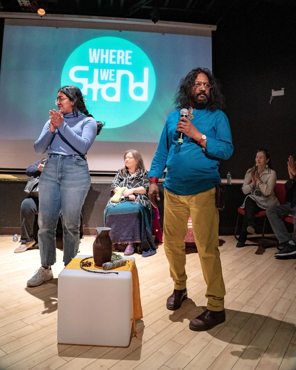 Two people standing front of an audience with Where We Stand logo in the background.
