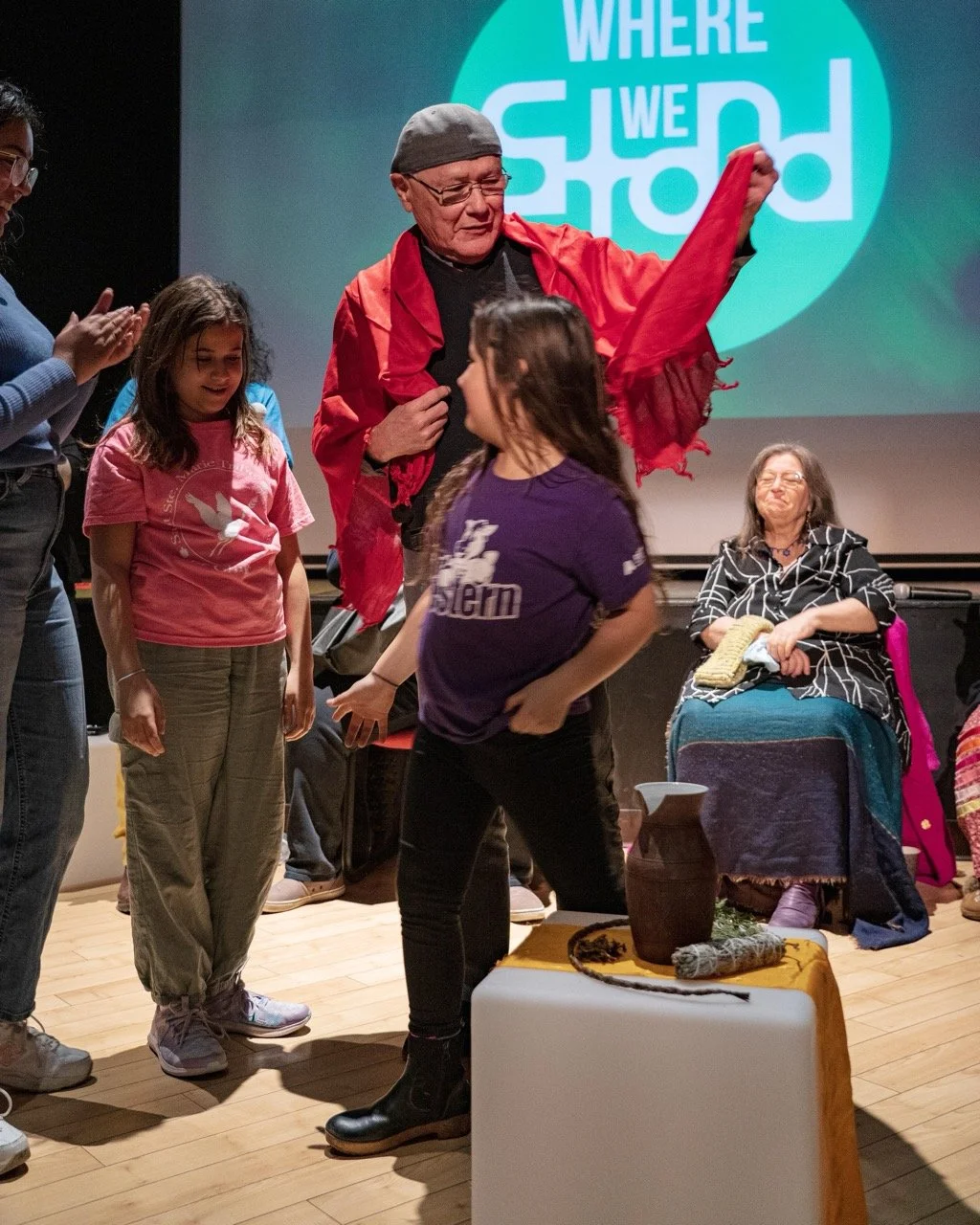 Three people are standing front of the Where We Stand logo and an audience is sitting around them.