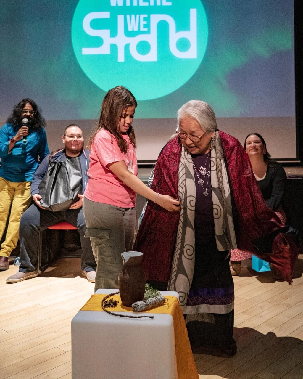 Two people are standing front of the Where We Stand logo and an audience is sitting around them.