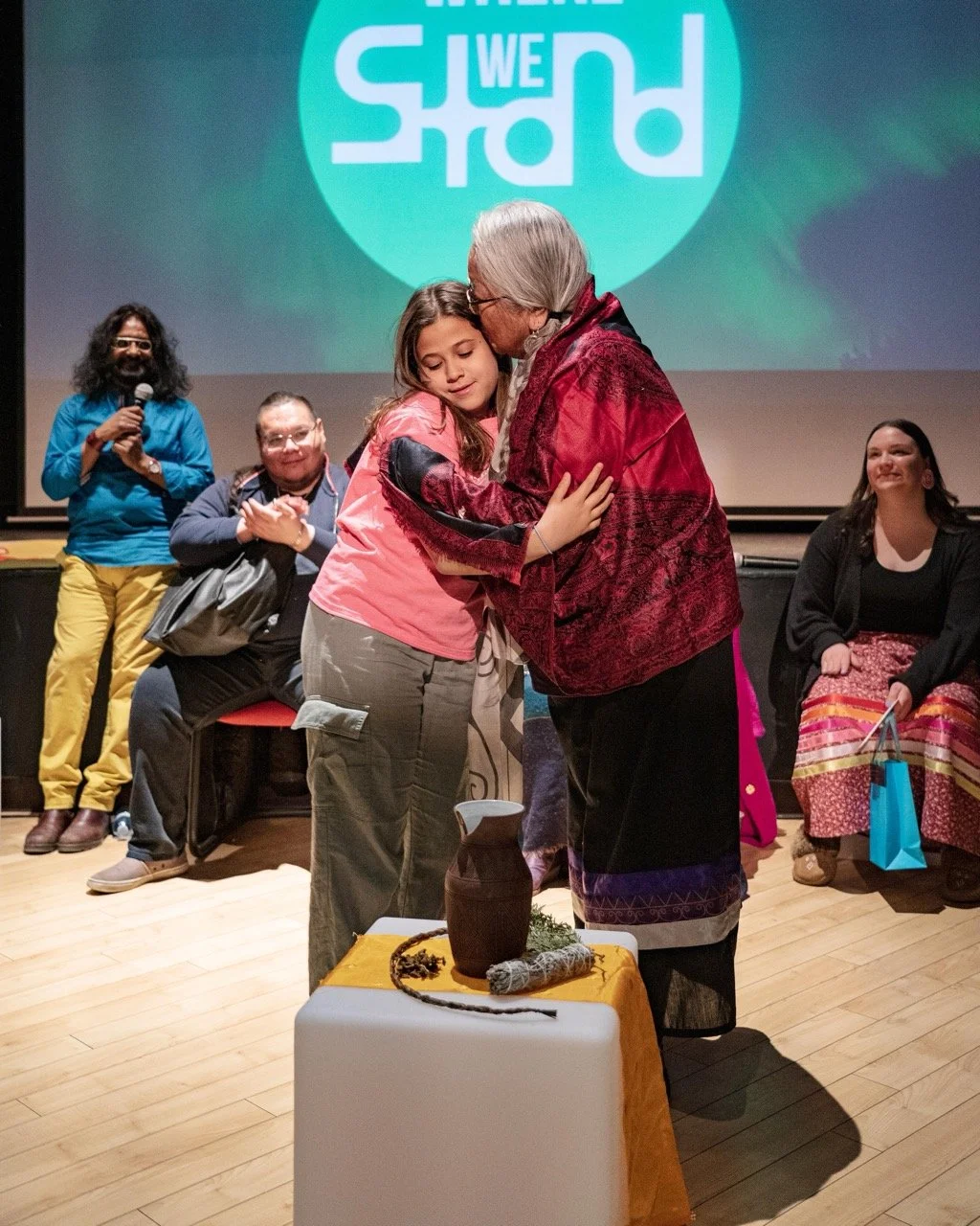 Two people are hugging front of the Where We Stand logo and an audience is sitting around them.