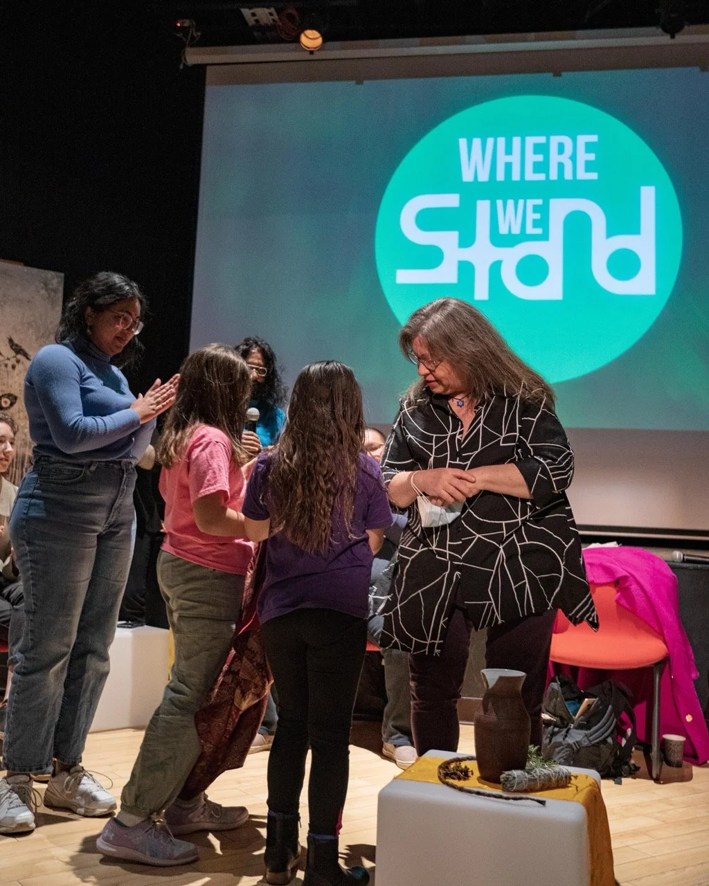 A group of people are standing front of the Where We Stand logo and an audience is sitting around them.
