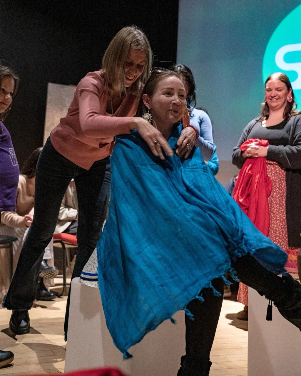One person sitting with a blue scarf on and an other person is standing behind them.