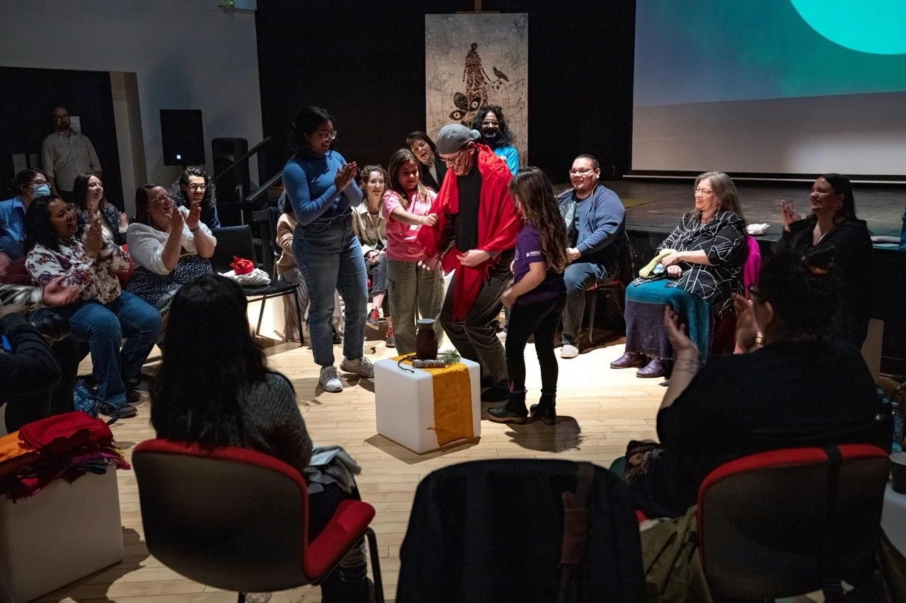 Wide angle of a group of people standing in the middle of a circle, front of an audience.