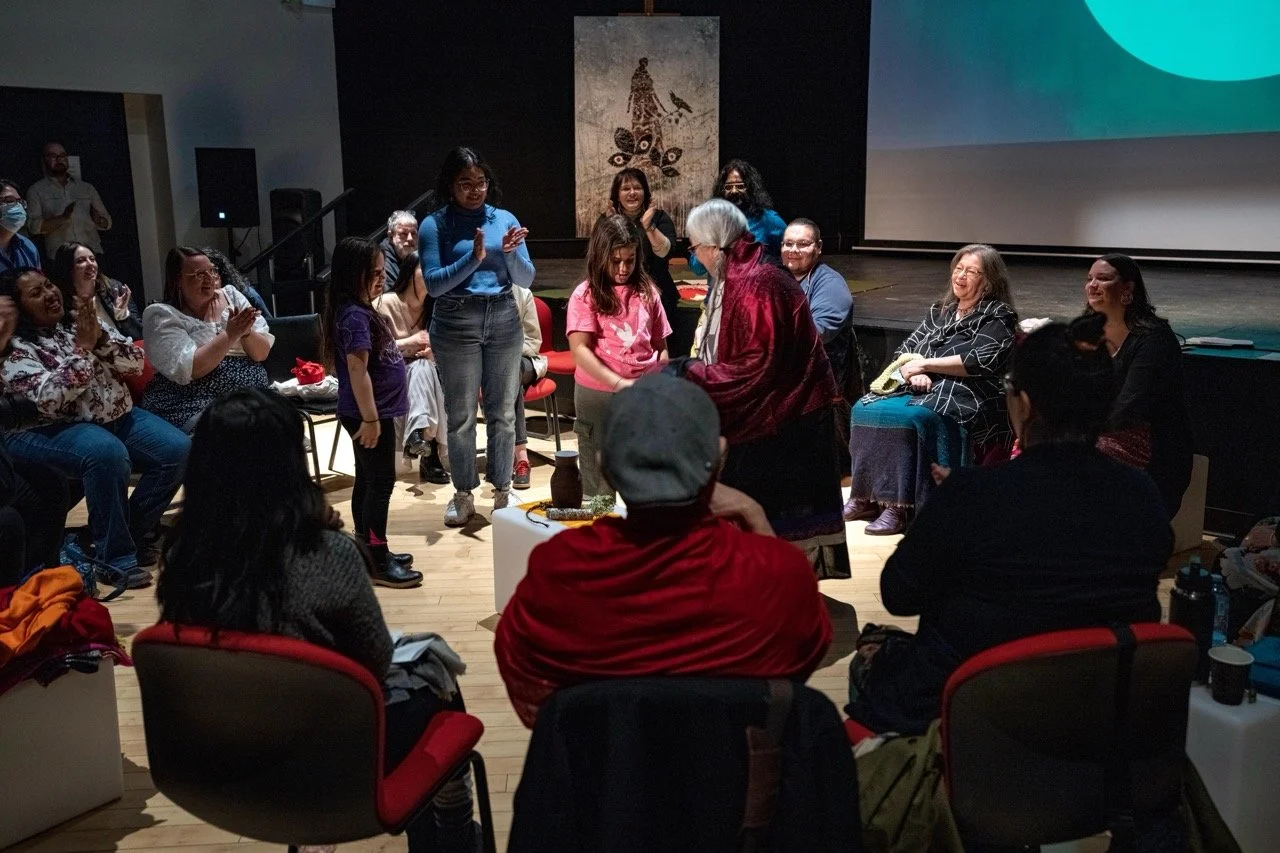 Wide angle of a group of people standing in the middle of a circle, front of an audience.