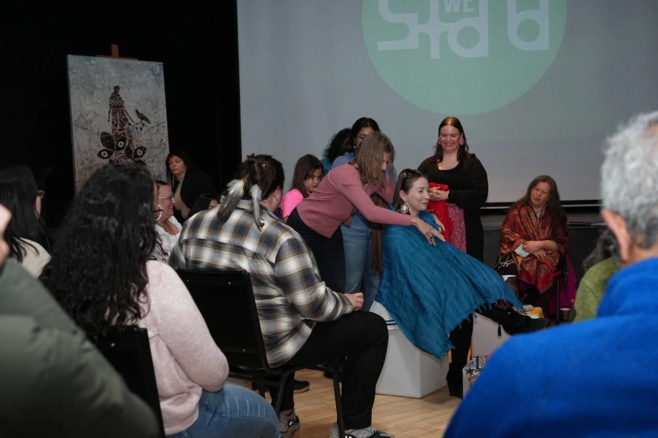 One person is putting a scarf on a person who is sitting front of them, with an audience around them.