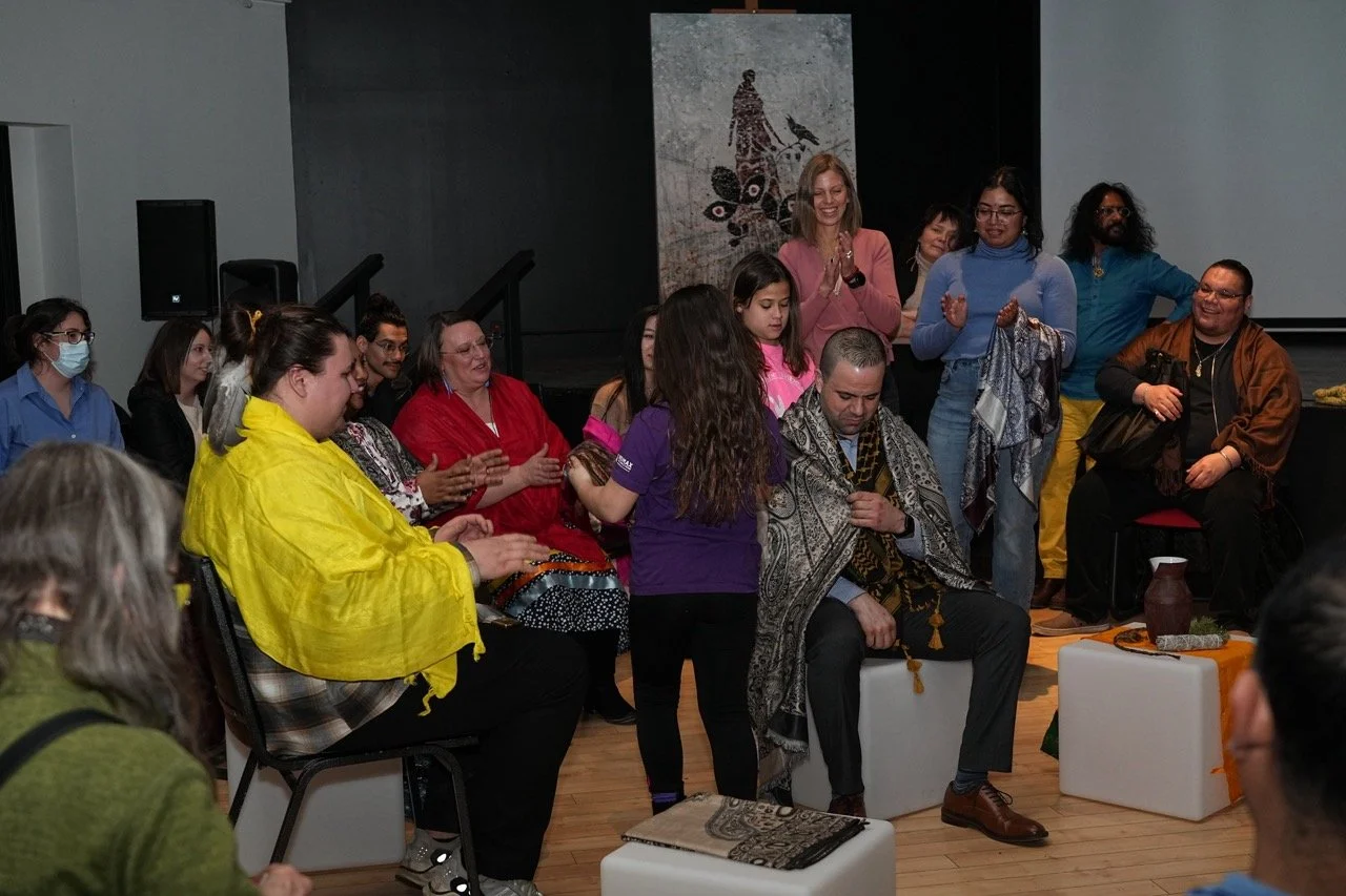 One person is putting a scarf on a person who is sitting front of them, with an audience around them.