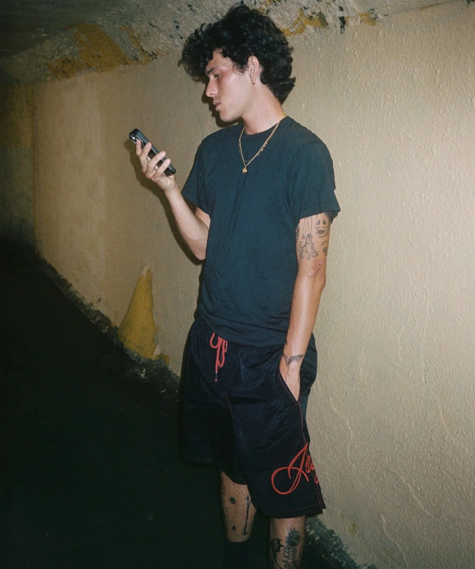 A young man with dark curly hair, wearing a black t-shirt, black shorts, tattoos on his arm and leg, gold necklaces, standing against a yellowish wall, looking at his phone.