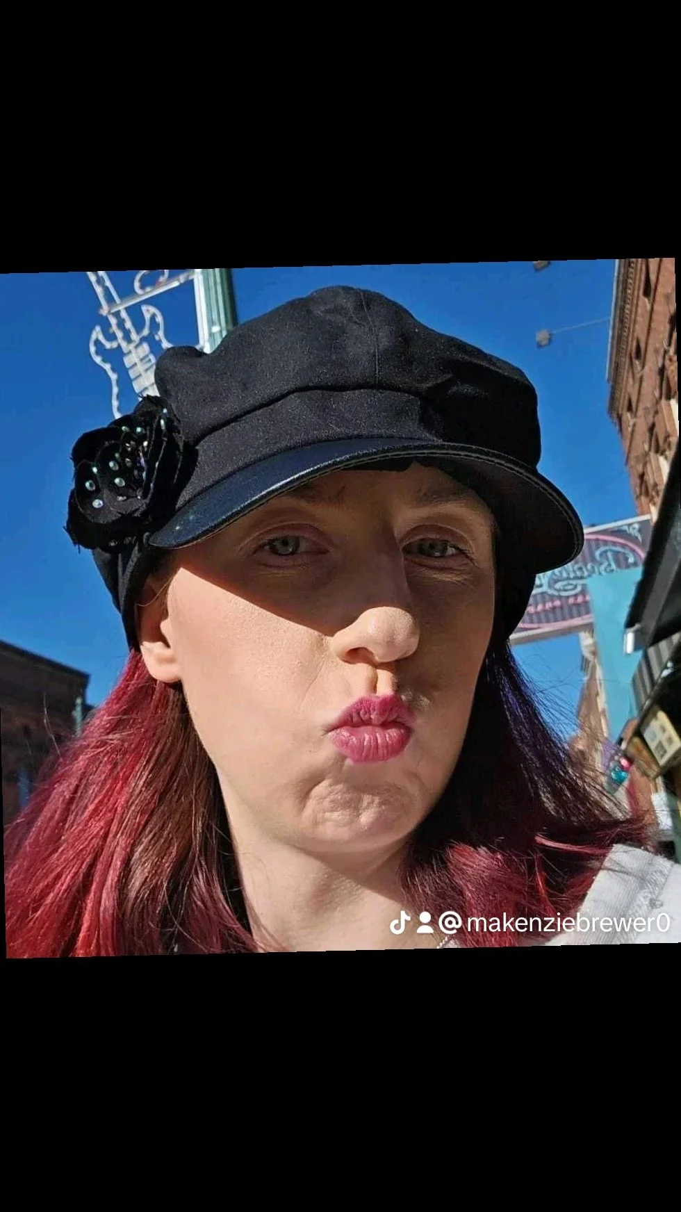 Kenzie (business owner) with a black hat and red hair posing outdoors under a blue sky.
