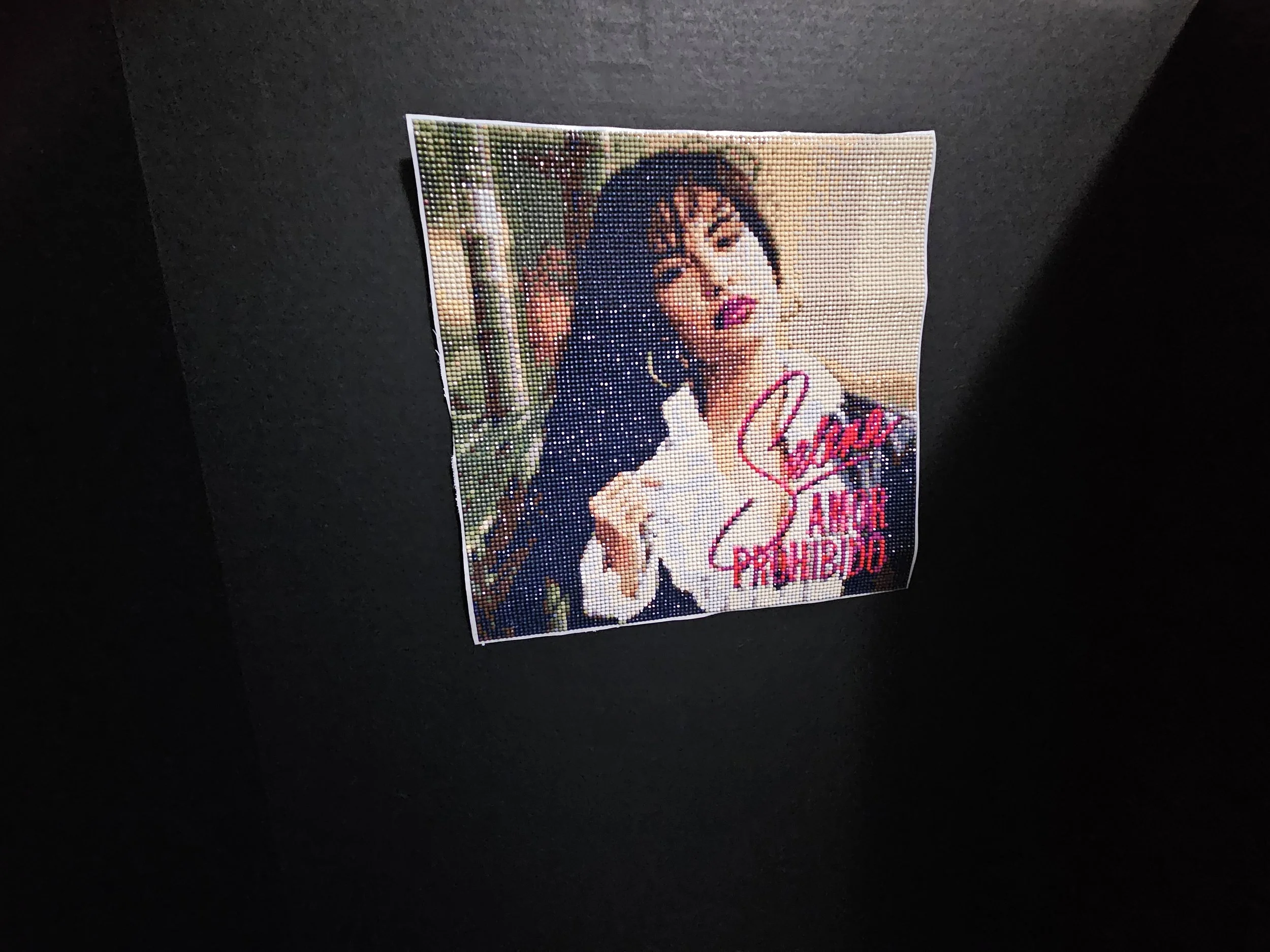 Mosaic artwork featuring Selena with dark hair, wearing a white and black outfit, set against a blurred outdoor background. Text reads "Amor Prohibido."