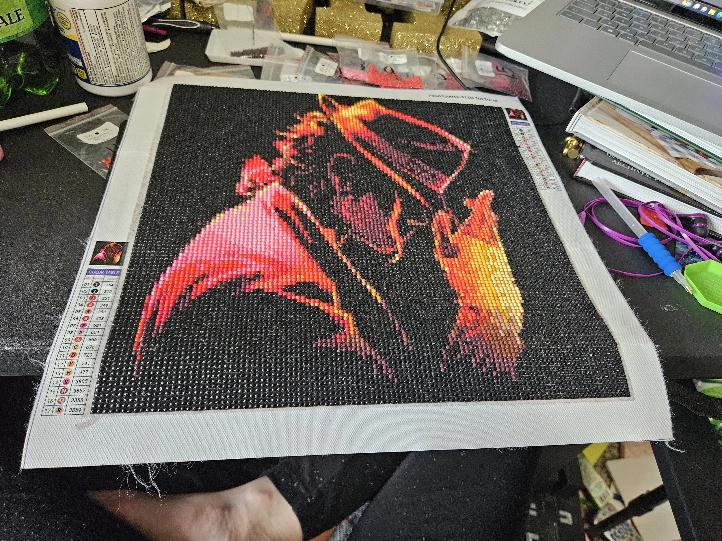 A pixel art depiction of Michael Jackson wearing a hat, in shades of red and orange, on a black background. The artwork is on a desk surrounded by craft materials and a laptop.