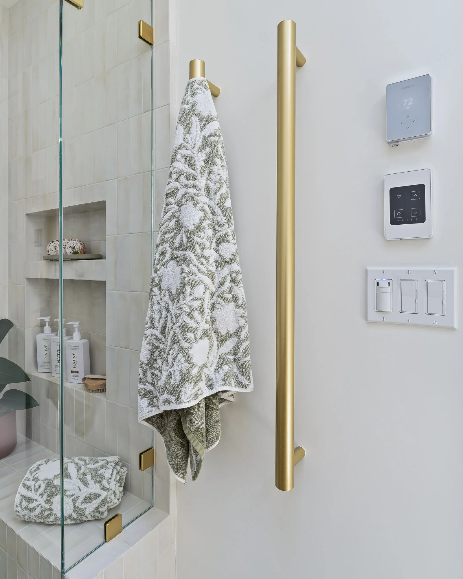 Brushed brass heated towel warmer installed beside a frameless glass shower in a modern bathroom remodel.