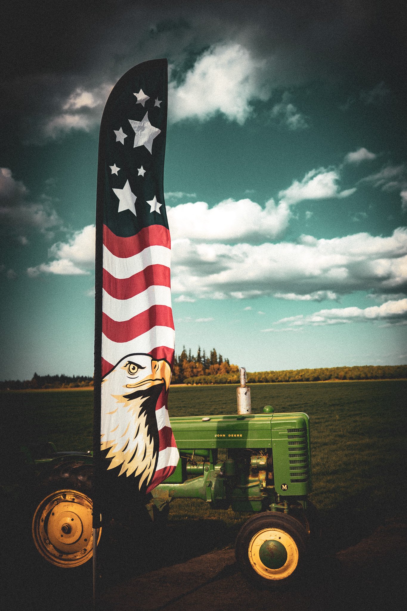 A green John Deere tractor parked on a field with a tall flag featuring an American eagle, red and white stripes, and stars, against a cloudy sky and distant trees.