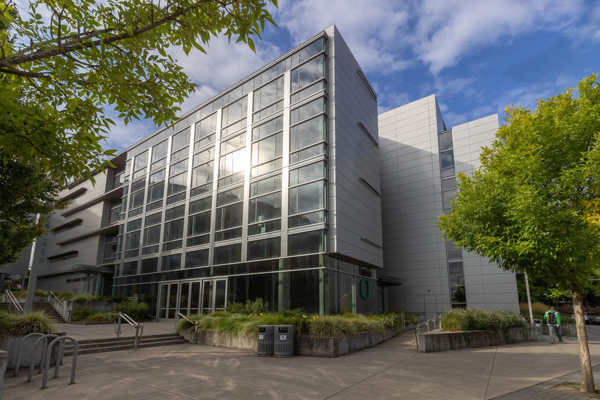Modern glass building with trees in foreground under a partly cloudy sky.