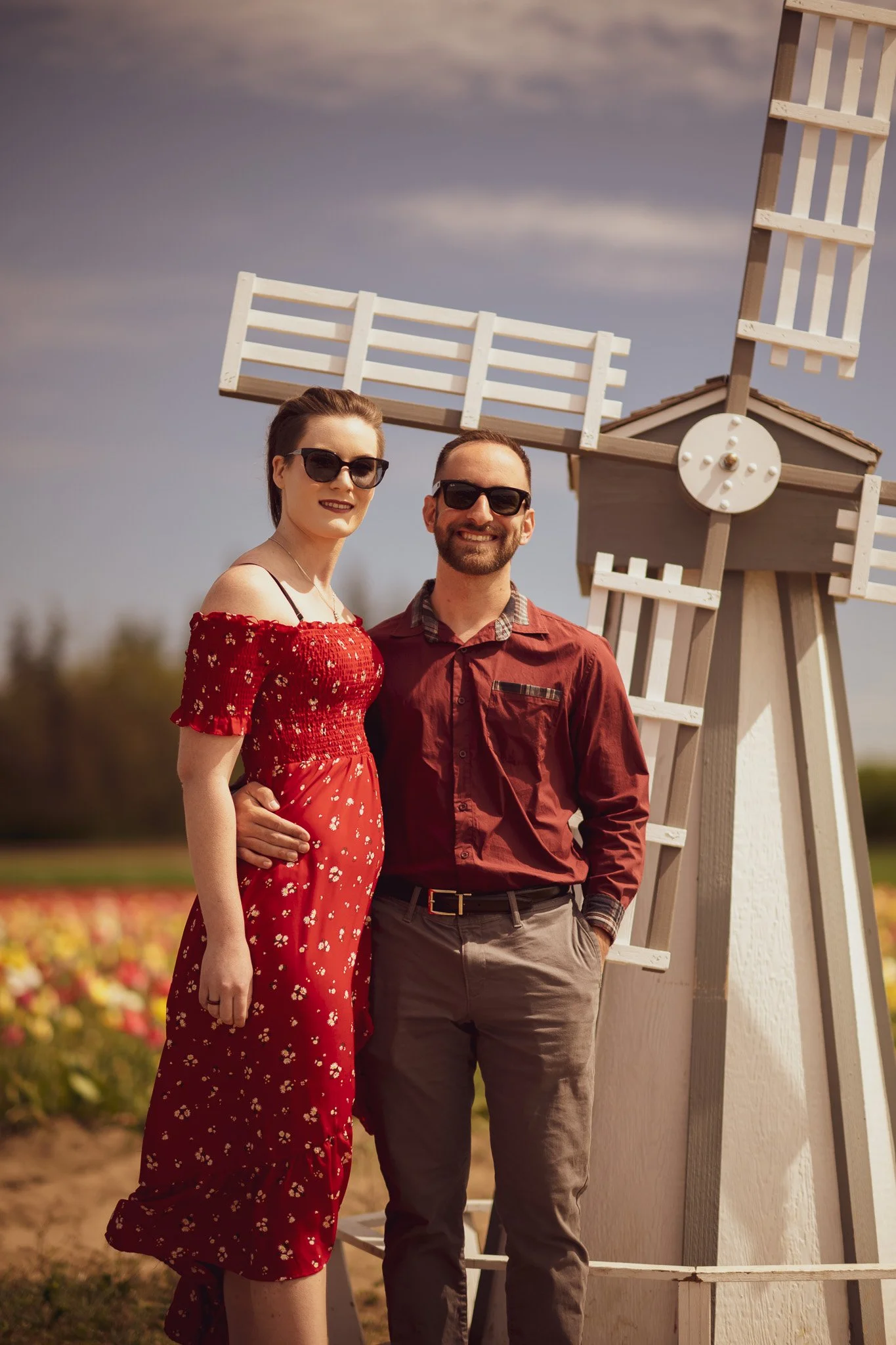 A couple standing in a field of colorful flowers next to a white wooden windmill, both wearing sunglasses and smiling.