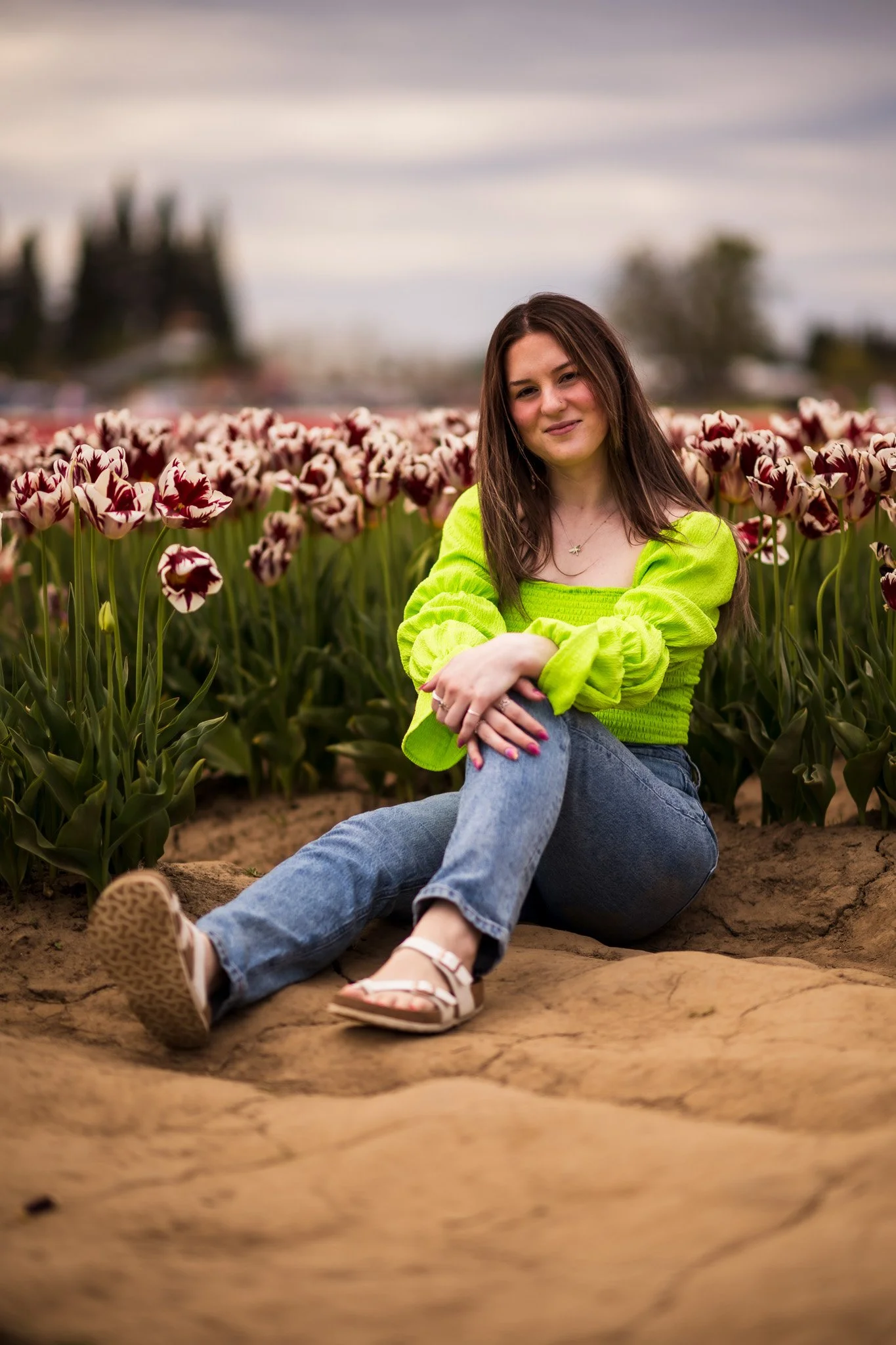 Young woman sitting on a rock in front of a tulip field, wearing a bright green top and jeans, with a cloudy sky in the background.