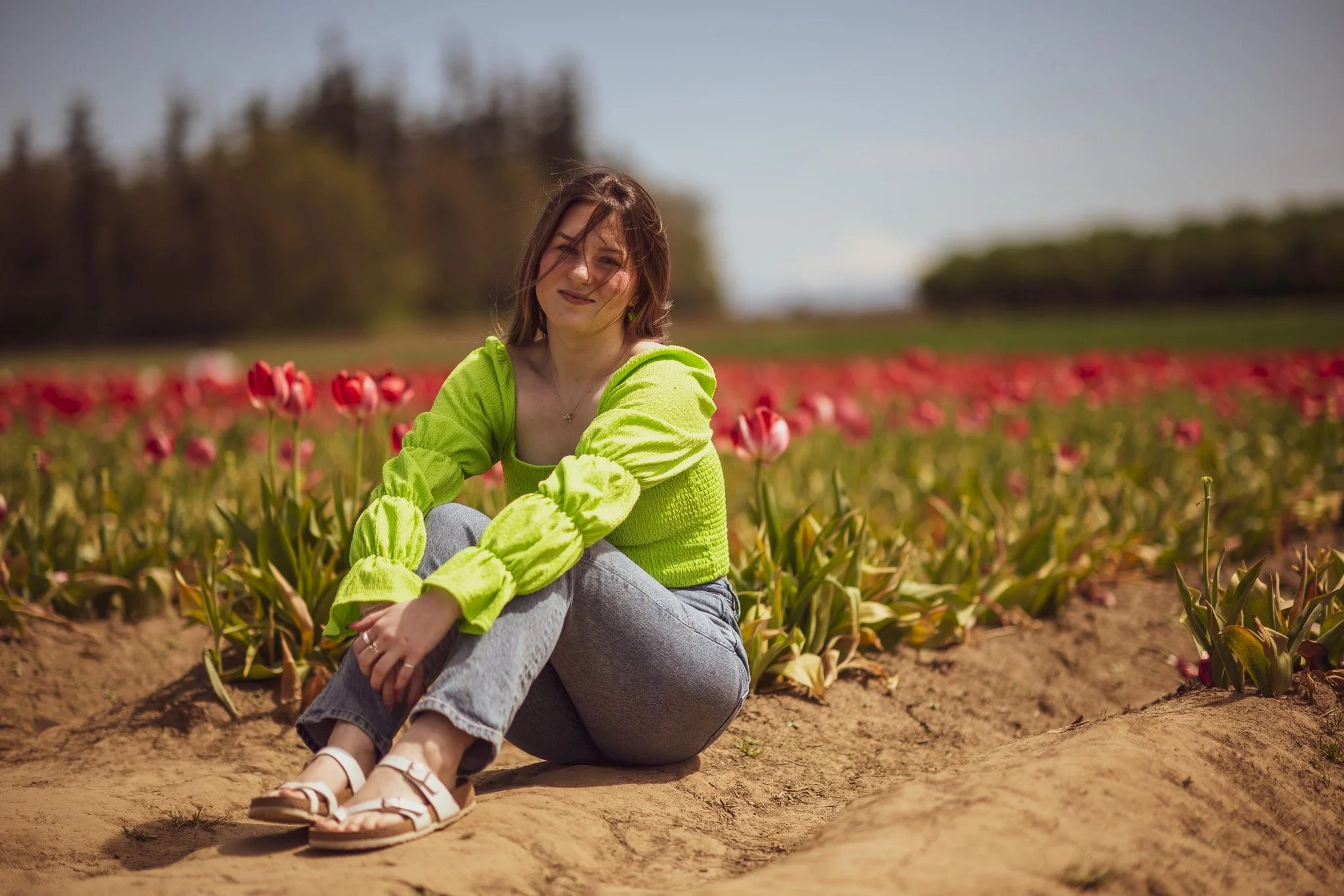 A young woman in a bright yellow-green sweater and jeans sitting on the ground among blooming tulips in a field on a sunny day.