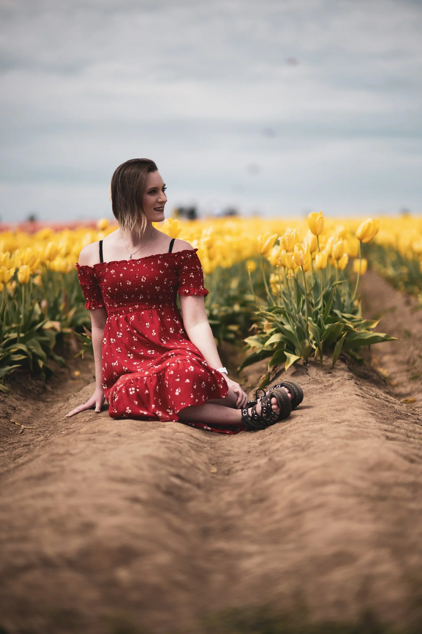 A woman in a red floral dress sitting on a dirt path between yellow tulip fields holding her legs, with a cloudy sky above.