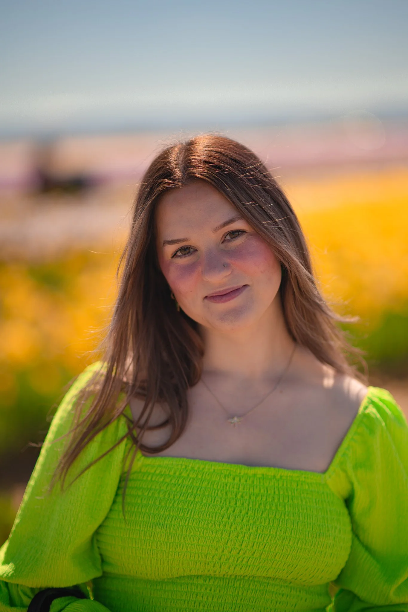 A young woman with long brown hair, a nose piercing, and a necklace, wearing a bright green top, standing outdoors with blurred yellow flowers in the background.