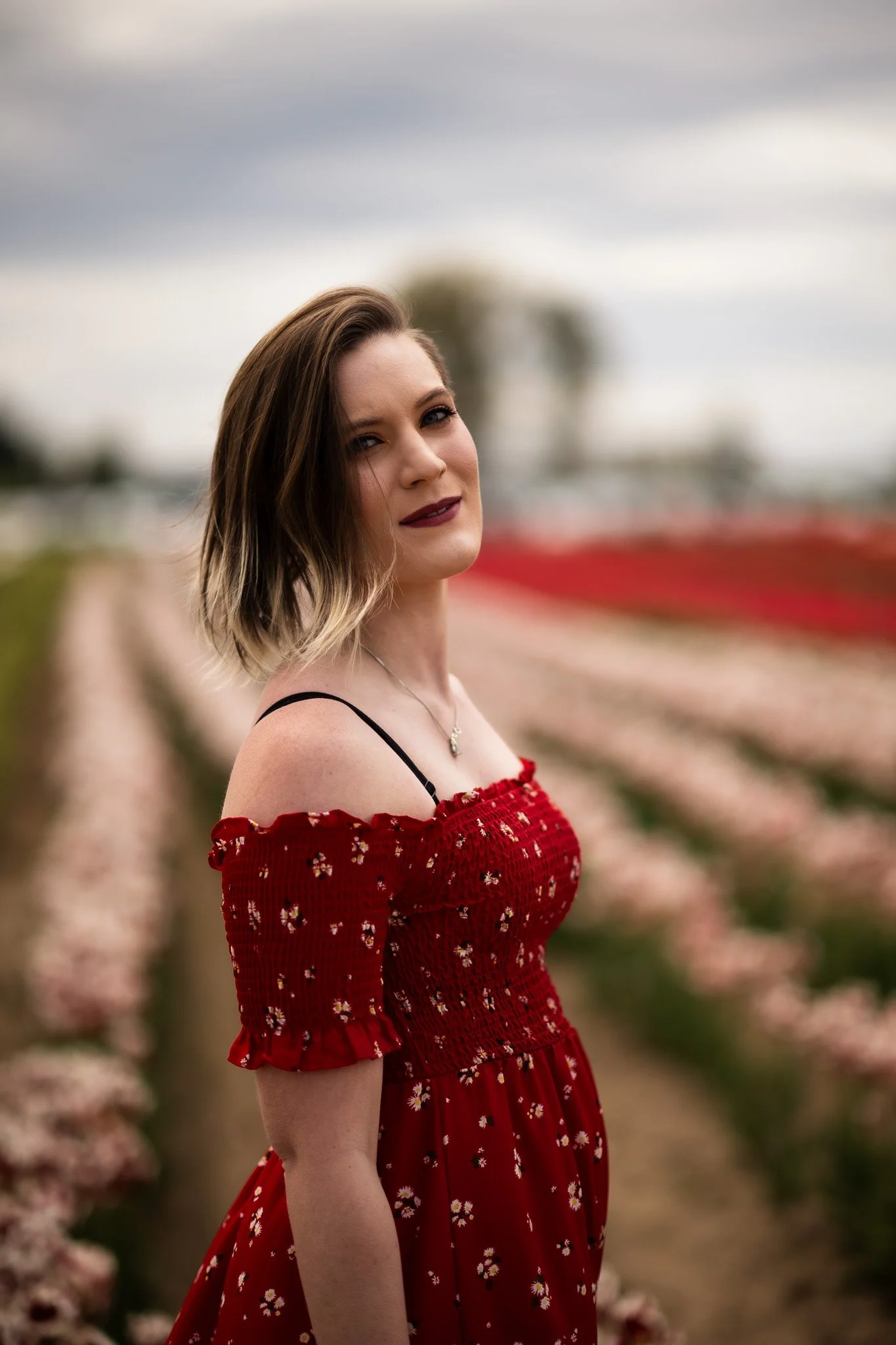 A woman with shoulder-length brown hair styled with blonde tips, wearing a red off-the-shoulder dress with small floral patterns, standing in a field of pink flowers under an overcast sky.