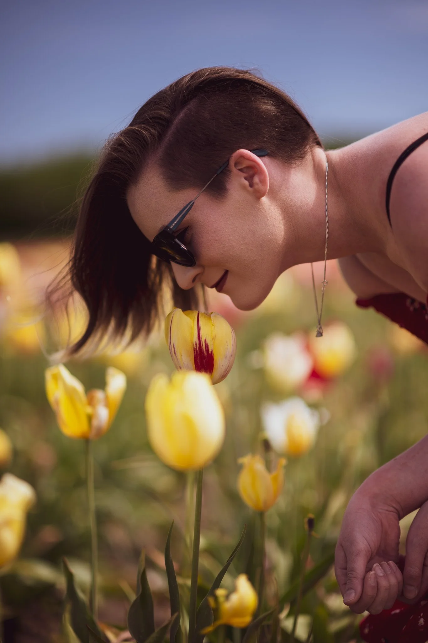 A woman with short brown hair, sunglasses, and a necklace is smelling yellow and red tulips in a field on a sunny day.