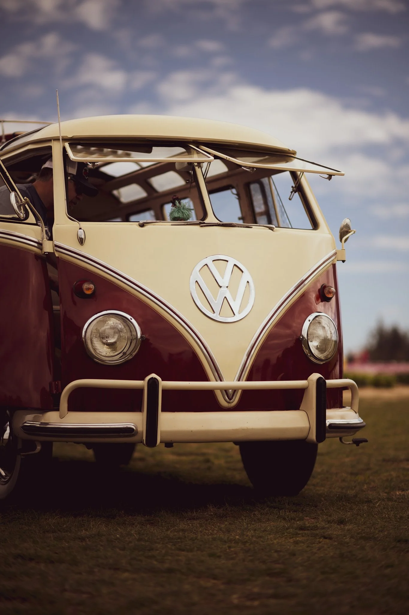A vintage red and cream Volkswagen Type 2 van parked outdoors on grass, with a partly cloudy sky in the background.