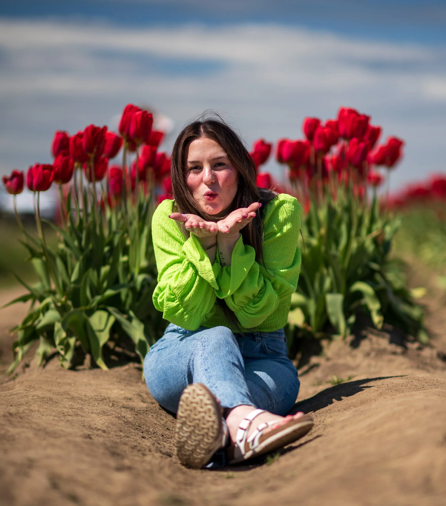 A woman blowing a kiss towards the camera while sitting on the ground in front of a row of red tulips in a field.