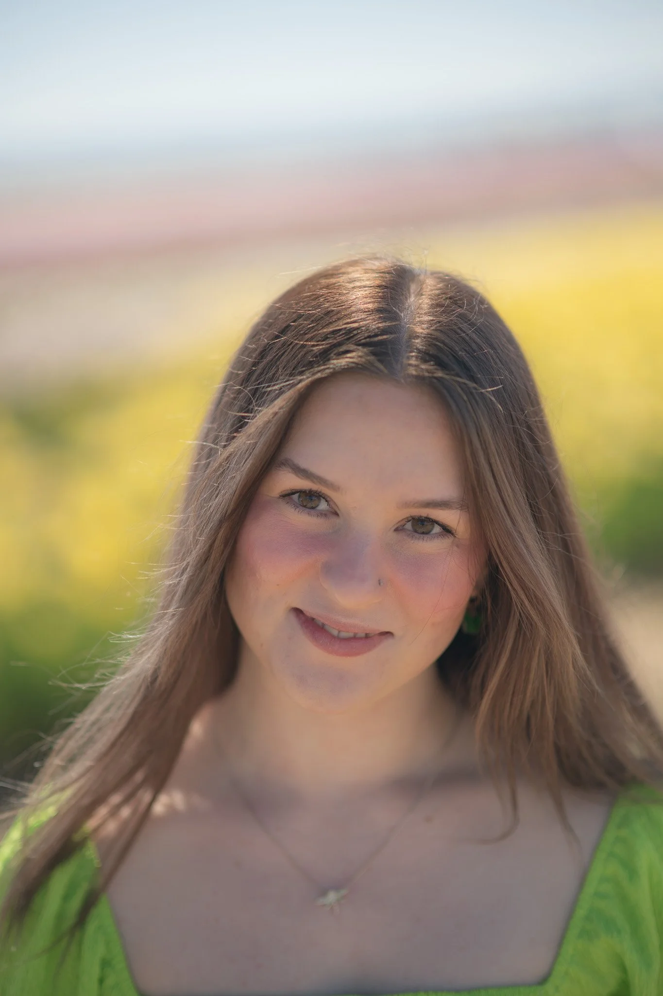 A young woman with long brown hair, smiling, outdoors in bright sunlight, wearing a green top and a necklace with a star pendant.