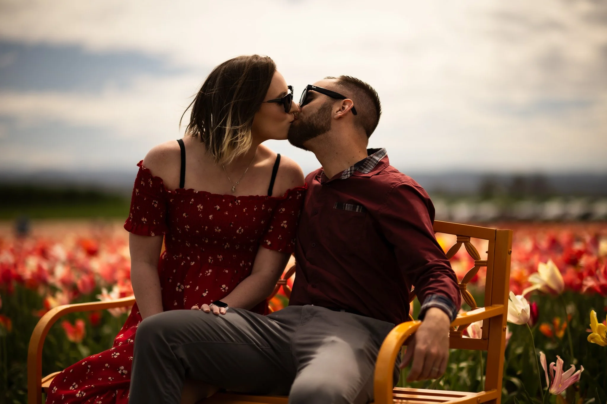 A couple sharing a kiss while sitting on a wooden bench in a field of tulips during sunset.