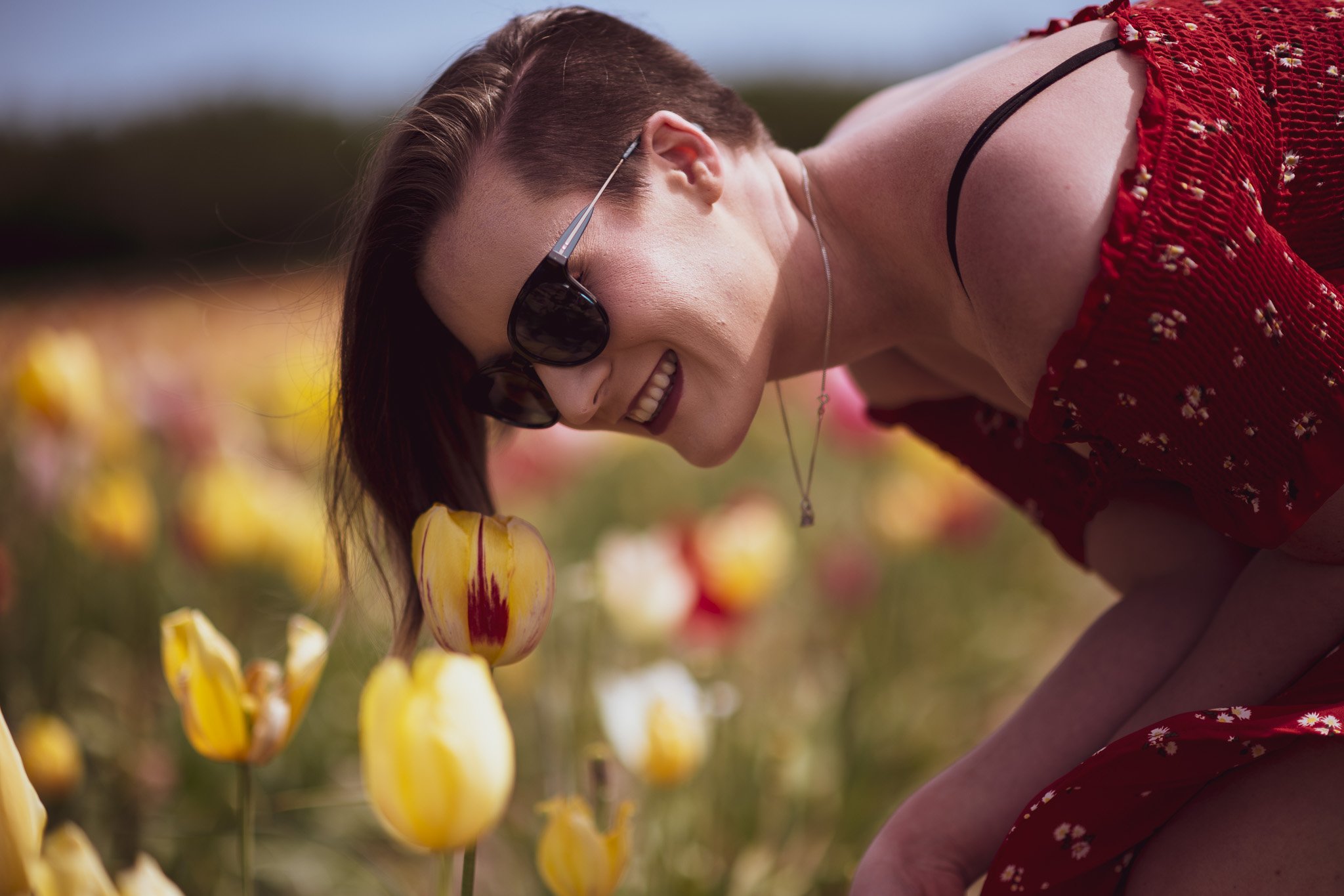 A woman in sunglasses smiling while bending over yellow and red tulips in a field.