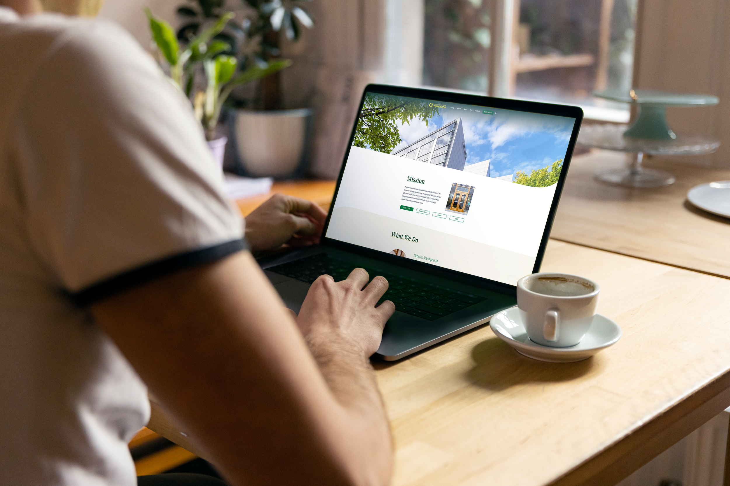 Person working on a laptop with a website about a mission project displayed, sitting at a wooden table with a cup of coffee, in a room with a large window, potted plant, and cake stand in the background.