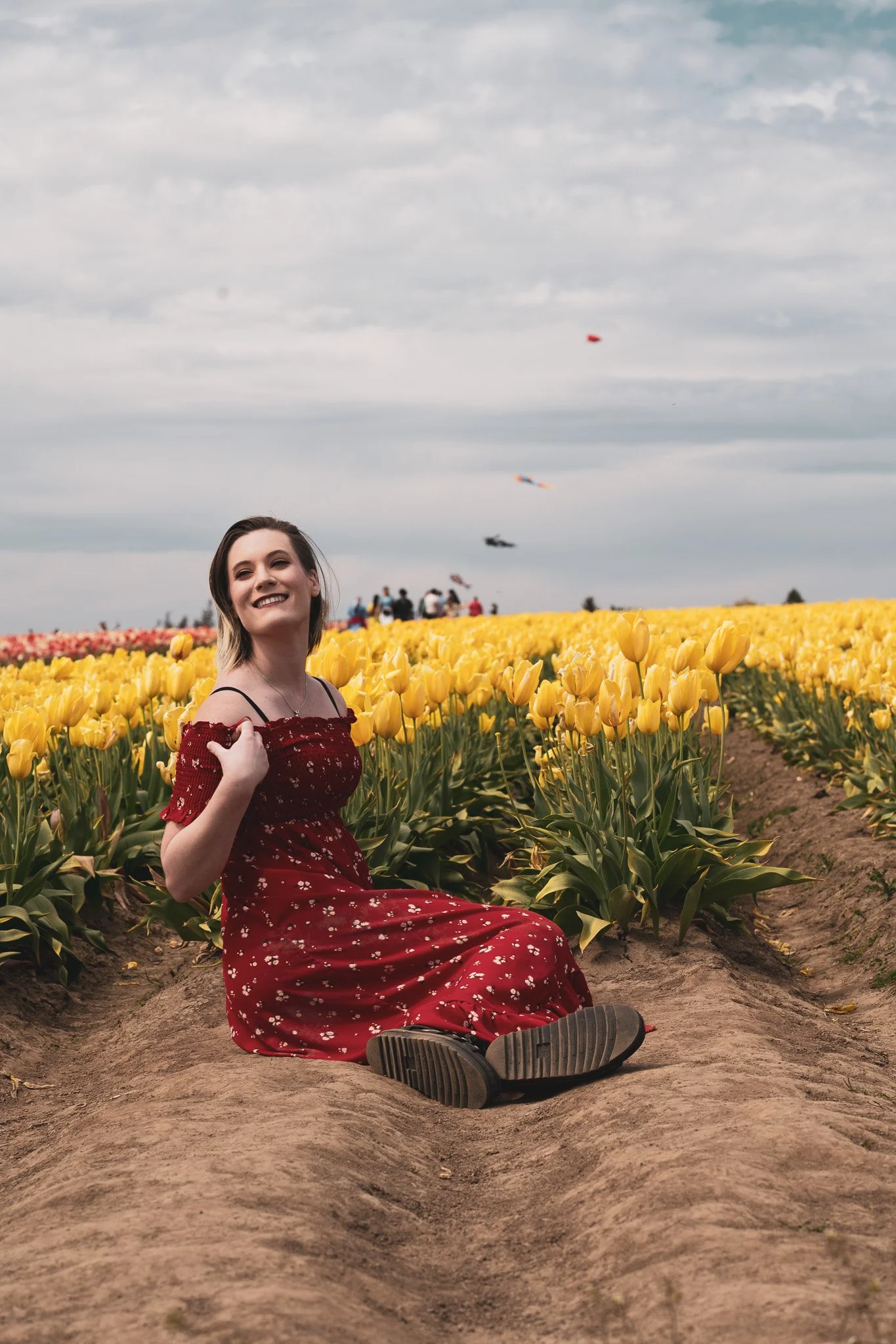 A woman sitting on the ground between rows of yellow tulips in a field, smiling and enjoying the day under a partly cloudy sky, with kites flying in the background.