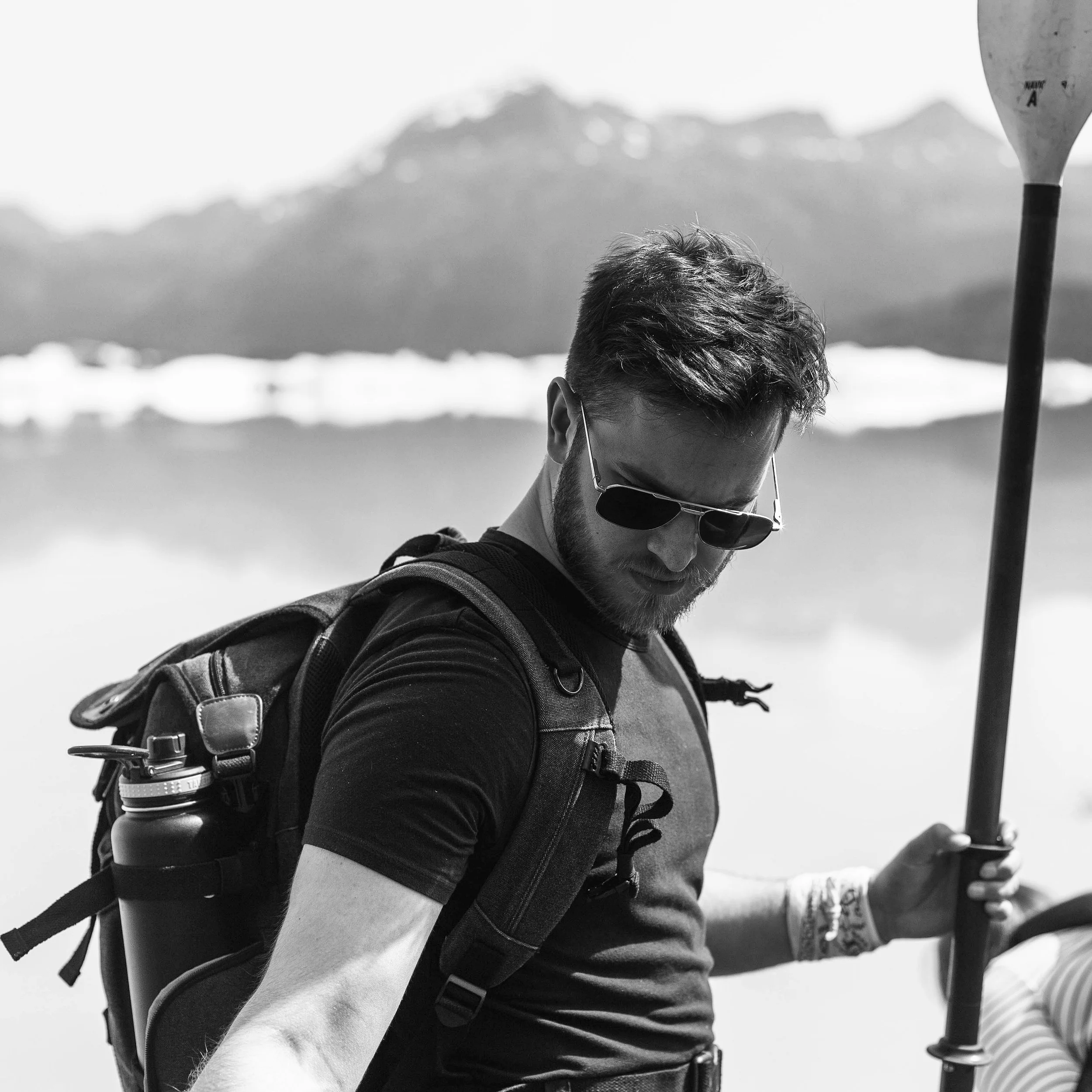 Black and white photo of a person with sunglasses holding a paddle, wearing a backpack, standing outdoors with a mountainous background.