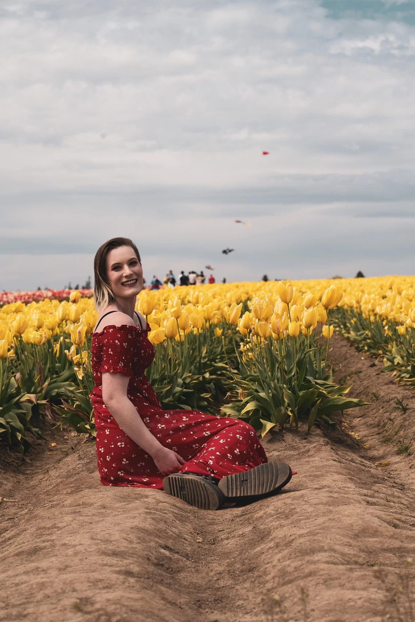 A woman in a red floral dress sitting on the dirt path between tulip fields with kites flying in the cloudy sky.