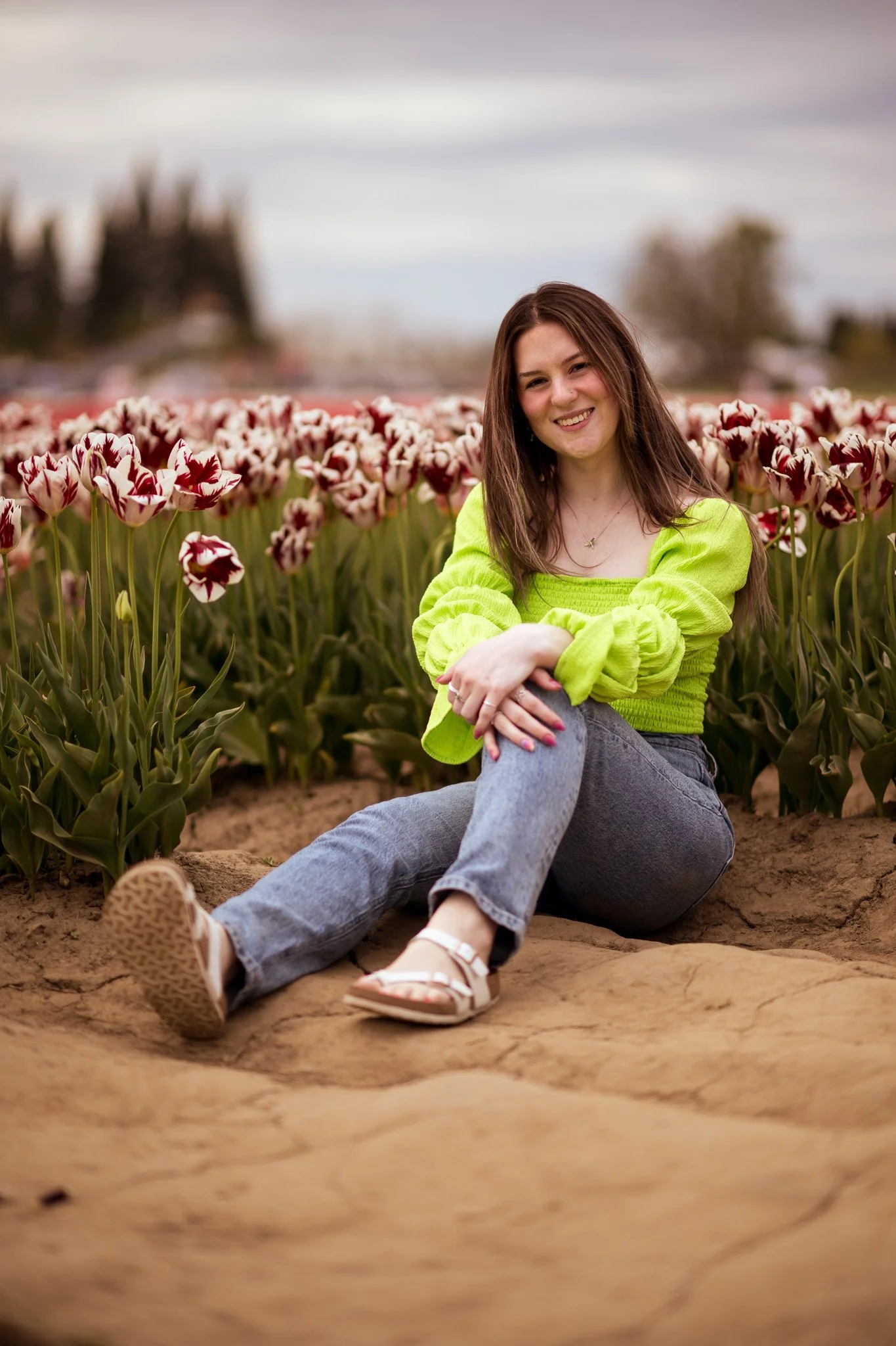 A young woman with long brown hair sitting on a large rock in front of a tulip field, smiling at the camera, wearing a bright green sweater, jeans, and sandals, with a blurred background of trees and cloudy sky.