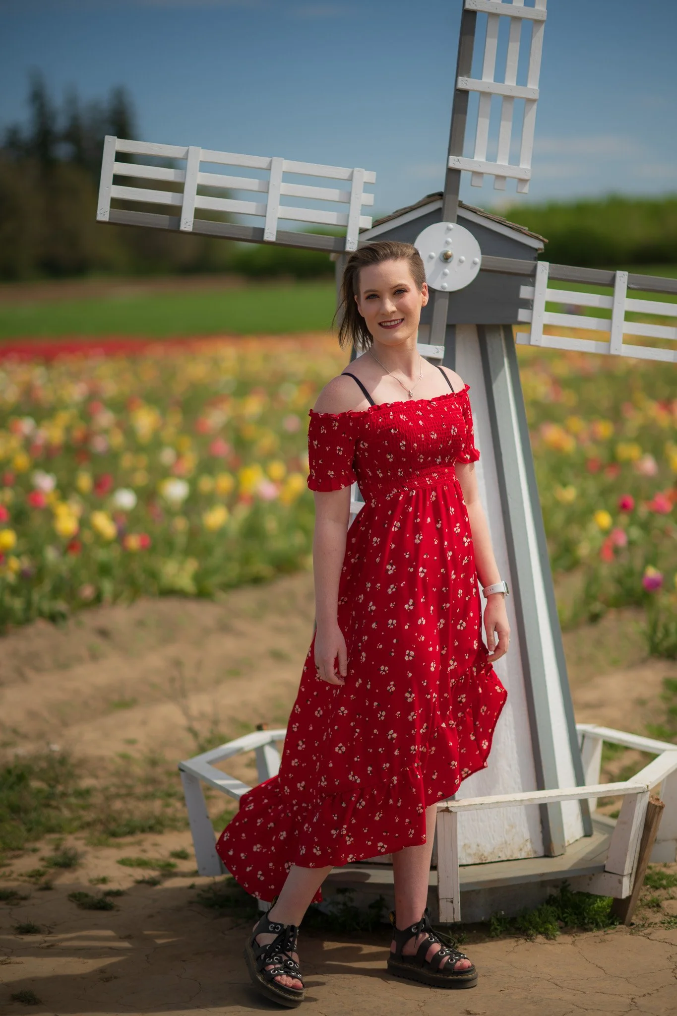A woman in a red floral dress standing next to a white windmill in a field of colorful flowers.