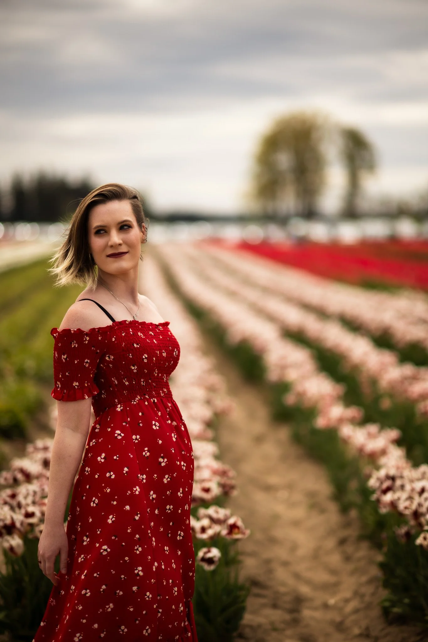 A woman in a red floral dress standing in a tulip field with rows of tulips extending behind her and a blurred background of trees and cloudy sky.