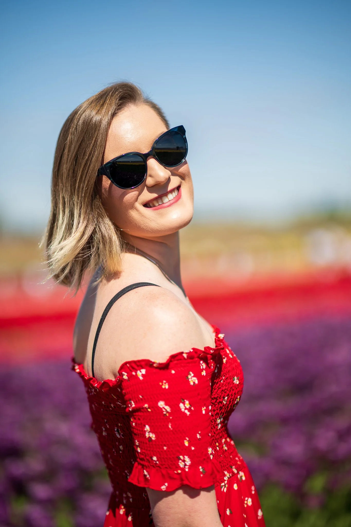 A woman smiling and wearing sunglasses in a field of colorful flowers on a sunny day.