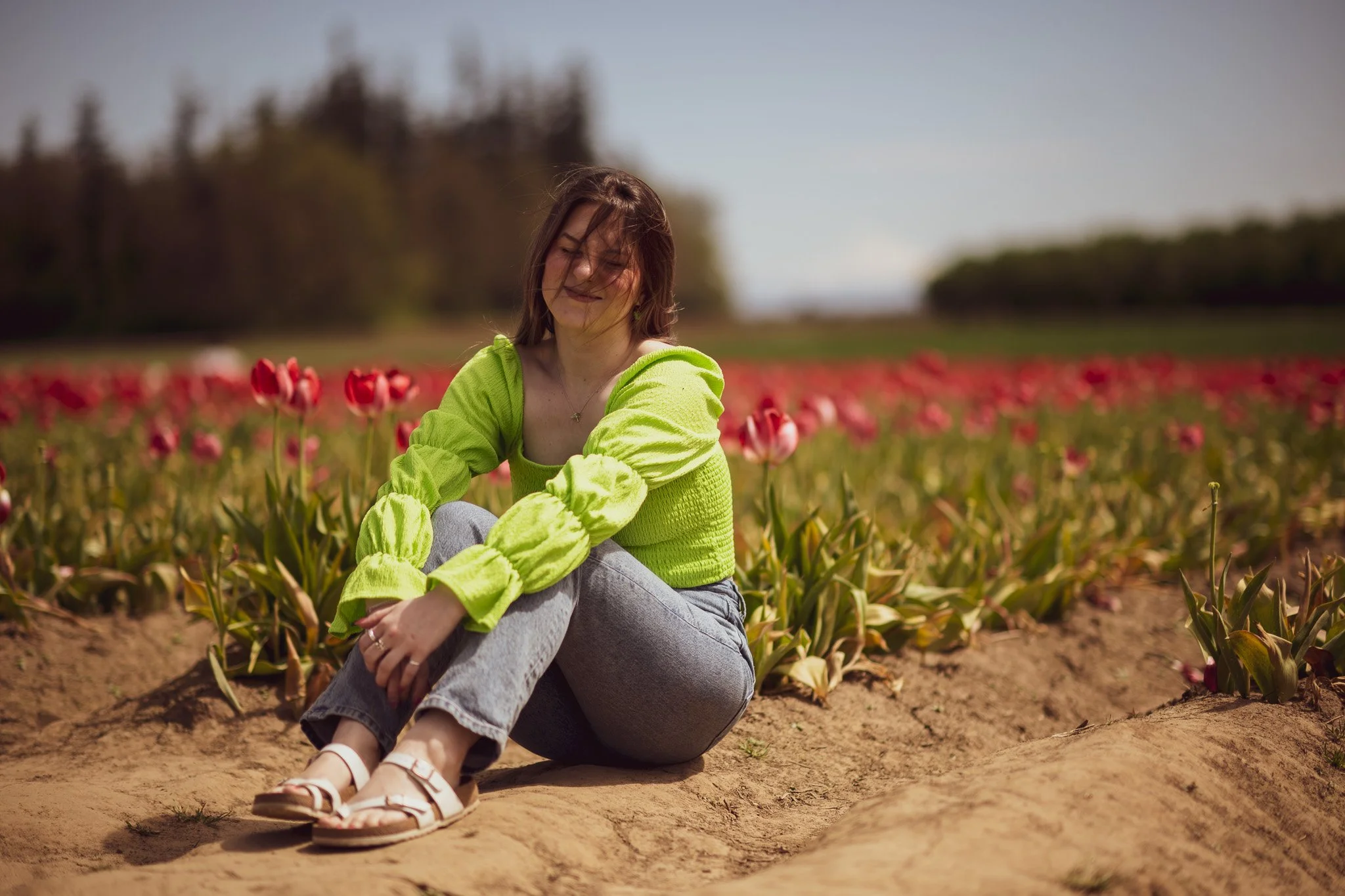 A woman sitting on the ground next to pink tulips in a field, smiling with closed eyes, wearing a lime green sweater and gray jeans.