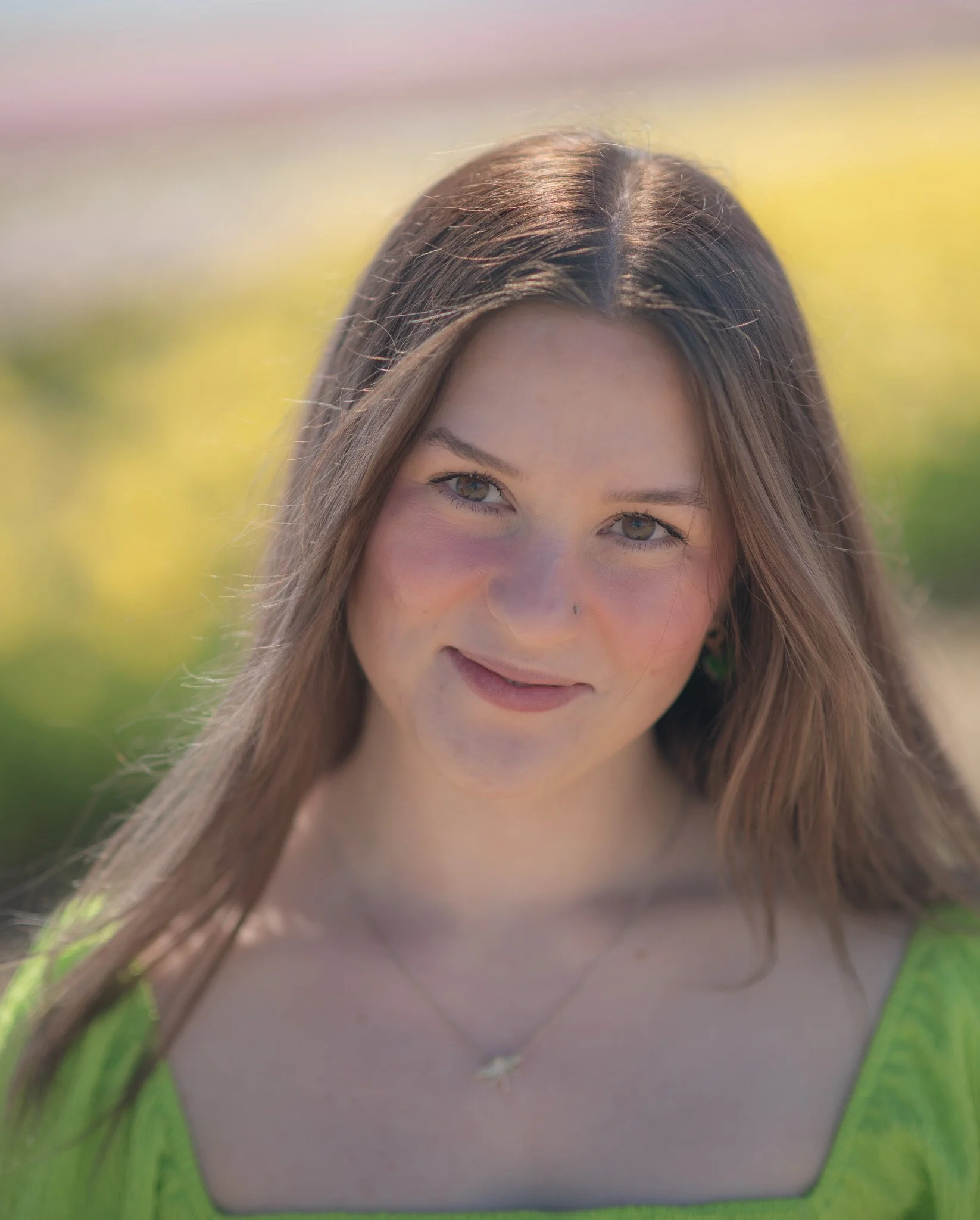 Close-up of a smiling young woman with long brown hair, wearing a green top and a necklace, outdoors with blurred greenery in the background.