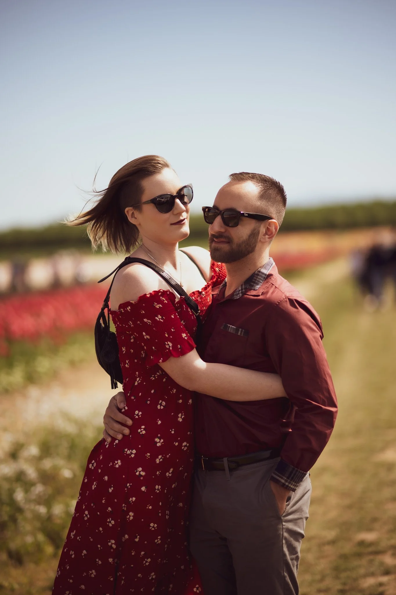 A couple in sunglasses embracing outdoors during daytime, with a field of colorful flowers in the background.