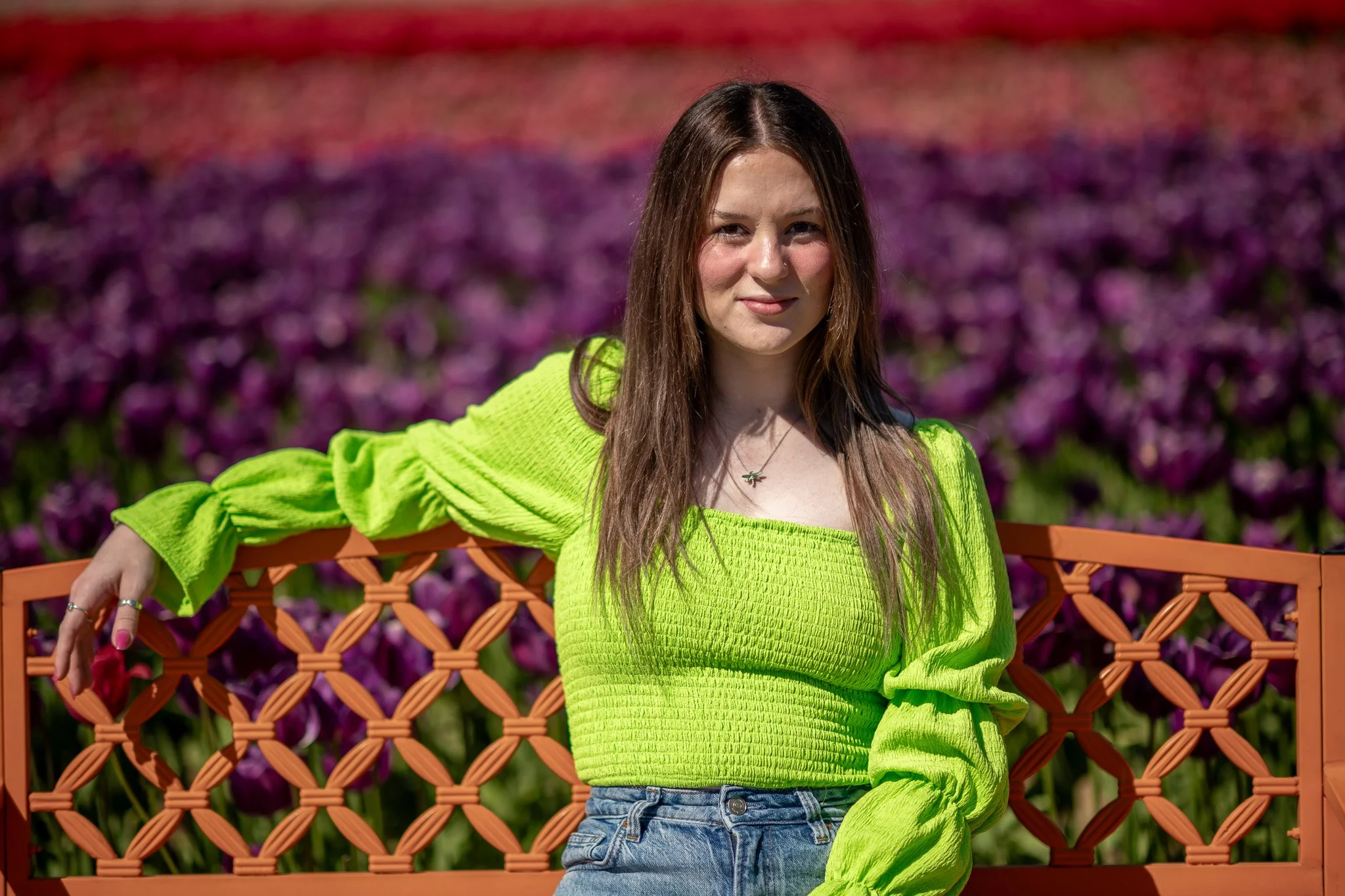 Young woman sitting on an orange bench with purple flowers in the background, wearing a bright green long-sleeve top and blue jeans.